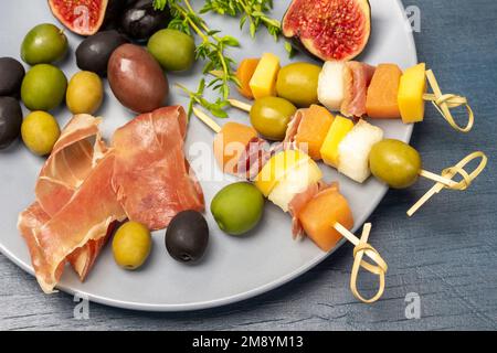 Spiedini con olive, formaggio e melone su un piatto grigio. Prosciutto, olive, fichi e rametti di timo. Giacitura piatta. Sfondo blu. Foto Stock