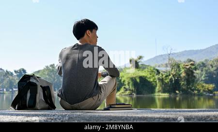 Bello e agghiacciante giovane studente asiatico maschio college in abiti casual rilassa seduti accanto al bellissimo stagno nel campus Park. vista posteriore Foto Stock