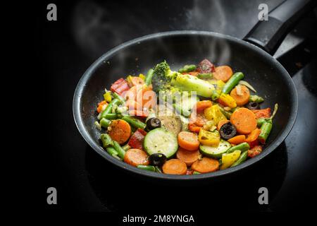 In una padella nera, mescolare le verdure saltate con carote, zucchine, broccoli, fagiolini, olive ed erbe aromatiche, Sano pasto vegetariano stile mediterraneo, copia Foto Stock