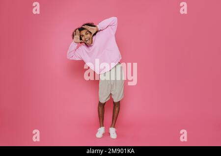 Giovane felice sorridente bell'elegante uomo latino riccio che tiene i capelli e mostra la lingua mentre si trova su sfondo rosa isolato Foto Stock