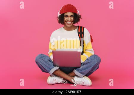 Giovane uomo caraibico in cuffie sorridente e utilizzando un computer portatile isolato su sfondo rosa Foto Stock