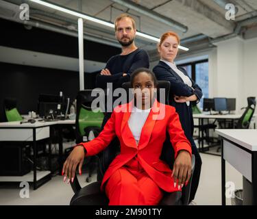 Donna caucasica dai capelli rossi, uomo caucasico bearded in piedi dietro una giovane donna afroamericana seduta in ufficio. Foto Stock