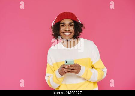 Giovane uomo caraibico in cuffie sorridente e utilizzando cellulare isolato su sfondo rosa Foto Stock