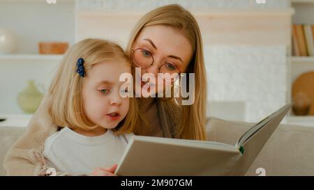 Madre caucasica negli occhiali con piccolo bambino cute figlia prescolare bambino ragazza lettura libro in soggiorno a casa mamma con bambino letto Foto Stock