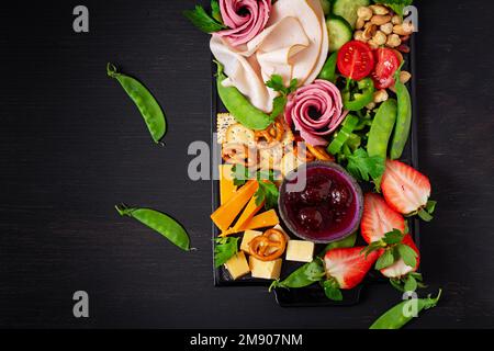 Antipasto piatto affettato freddo prosciutto, salame, cracker, fragole, verdure e formaggio piatto a bordo su sfondo scuro. Antipasti tavolo con Foto Stock