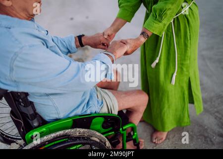 Primo piano di una donna anziana che tiene le mani sul marito in sedia a rotelle, godendo le vacanze insieme. Foto Stock