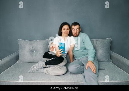 Coppia millenaria con disgusto espressione del volto perché reazione avversione guardando lo schermo dello smartphone seduto sul divano a casa. Concetto di emozioni umane Foto Stock