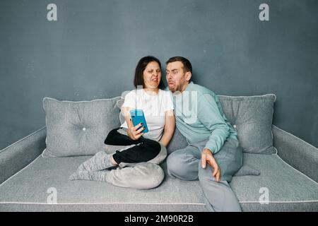Coppia millenaria con disgusto espressione del volto perché reazione avversione guardando lo schermo dello smartphone seduto sul divano a casa. Concetto di emozioni umane Foto Stock