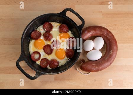 Vista dall'alto delle uova fritte con fette di pancetta. Salsiccia cruda e uova non rotte sono accanto alla padella su sfondo di legno. Foto Stock