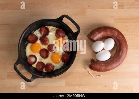 Vista dall'alto delle uova fritte con fette di salsiccia. Salsiccia cruda e uova non rotte sono accanto alla padella su sfondo di legno. Foto Stock