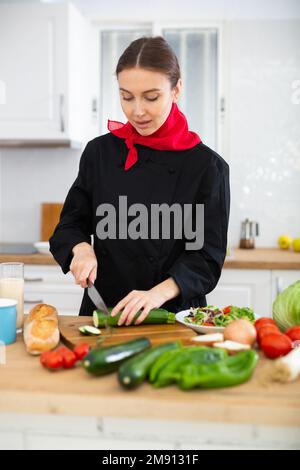 Sorridente cuoco femminile in uniforme nera che prepara insalata di verdure in cucina privata Foto Stock