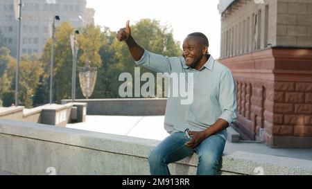 Amichevole medio-vecchio adulto maturo africano americano uomo d'affari imprenditore maschio seduto all'aperto in città in attesa di riunione data che agita la mano Foto Stock