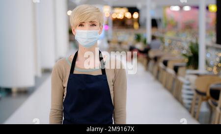 Adulto maturo di mezza età 40s donna in maschera medica ristorante cafe lavoratore guardando fotocamera incrocio braccia donna cameriera donna in grembiule lavoro Foto Stock