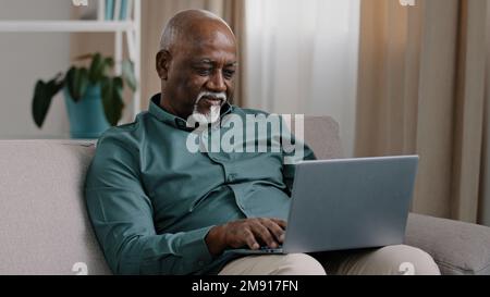 Vecchio calvo Africano americano uomo 60s senior maturo businessman utilizzare la tecnologia wireless facile per il lavoro comfort online shopping ecommerce a casa divano Foto Stock