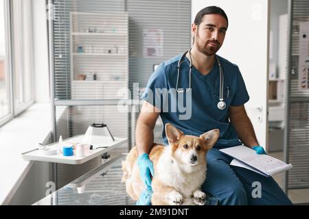 Giovane medico veterinario maschio fiducioso in uniforme e cane corgi malato seduto sul posto di lavoro in ufficio medico prima di esame Foto Stock
