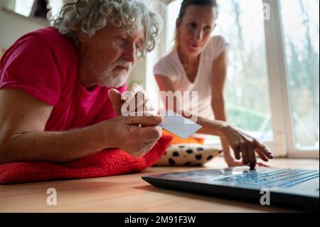 Giovane donna che insegna a un uomo anziano, suo padre, come usare un computer portatile per lo shopping online e l'istruzione. Entrambi sul pavimento a casa in li Foto Stock