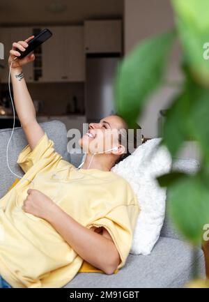 Giovane bruna sdraiata sul divano soggiorno e scattando selfie con il suo cellulare. Foto Stock