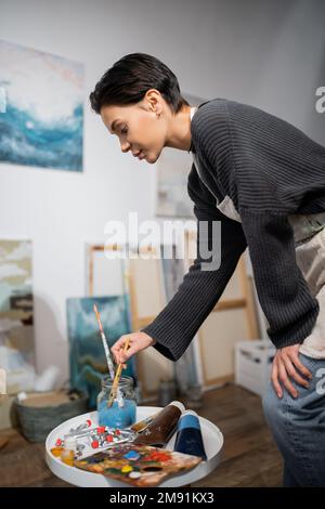 Vista laterale del giovane artista che prende il pennello dal vaso vicino alla tavolozza e le pitture in studio Foto Stock