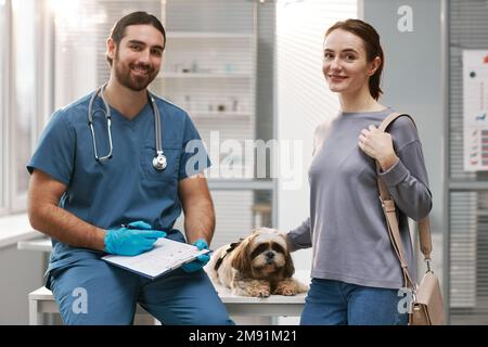 Giovane donna proprietaria di carino yorkshire terrier, il suo animale domestico e veterinario clinico in uniforme blu guardando la macchina fotografica in ufficio medico Foto Stock