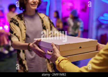 Primo piano di un giovane uomo sorridente che prende le scatole con la pizza passata dal liberayman in uniforme contro il gruppo di amici che hanno partito Foto Stock