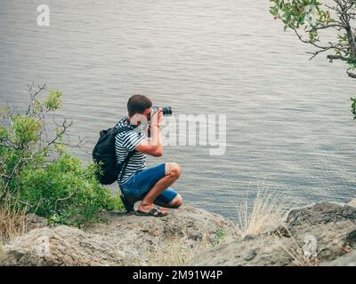 fotografo turista maschile che scatta foto seduto sul bordo di una scogliera mare Foto Stock