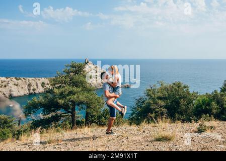 coppia affettuosa che abbraccia in estate il mare in montagna nel pomeriggio. Il ragazzo tiene la ragazza sulle mani, sono felici e sorridenti Foto Stock