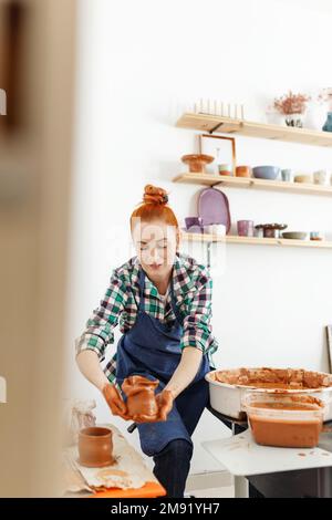 Allegra scultore femminile mentre lei fa pentola di argilla su ruota di ceramica. Idea di piccola impresa e imprenditorialità. Hobby domestico, intrattenimento e leisur Foto Stock