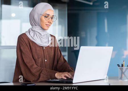 Donna d'affari sorridente e sognante che lavora all'interno dell'ufficio con un computer portatile, una donna in hijab e una lavoratrice in ufficio con occhiali felici e soddisfatti del lavoro seduto alla scrivania. Foto Stock