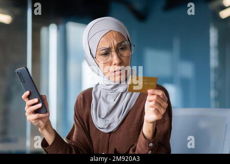 Donna sconvolta e delusa in hijab in ufficio cercando di fare l'acquisto in negozio online, donna d'affari in possesso di smartphone e carta di credito bancario, ricevuto errore di trasferimento di denaro. Foto Stock