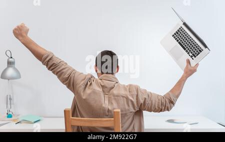 Un giovane uomo irriconoscibile con notebook festeggia con successo a braccia sollevate al lavoro in ufficio a casa. Foto Stock