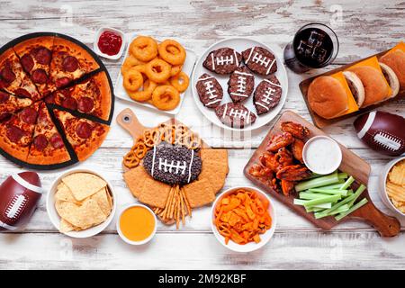 Superbowl o calcio tavolo alimentare a tema scena. Pizza, hamburger, ali di pollo, spuntini e contorni. Vista dall'alto su uno sfondo di legno bianco. Foto Stock