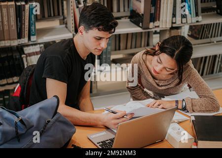 Gli studenti hanno concentrato gli studenti in abiti casual seduti al tavolo di legno con un computer portatile e lavorando insieme al progetto universitario mentre studiavano in biblioteca Foto Stock