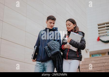 Da sotto giovani studenti positivi in abiti caldi con zaini e computer portatili sorridenti e salendo le scale mentre si va all'università Foto Stock