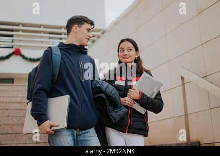 Da sotto giovani studenti positivi in abiti caldi con zaini e computer portatili sorridenti e salendo le scale mentre si va all'università Foto Stock