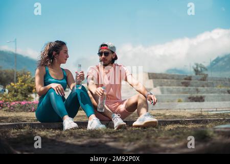 Corpo pieno di uomo in rosa sportswear e donna in verde activewear seduta a terra con bottiglie d'acqua durante la conversazione Foto Stock