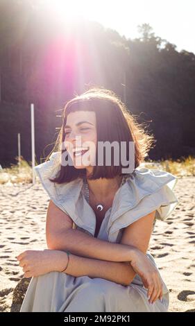 Giovane ragazza turista allegra con capelli scuri in abito elegante sorridente luminoso e guardando lontano mentre riposa sulla spiaggia di sabbia nelle giornate di sole Foto Stock