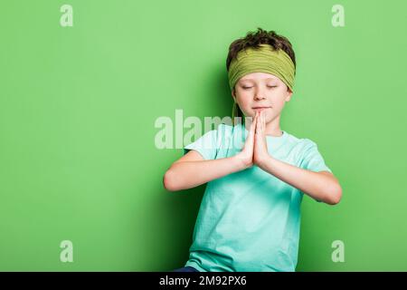 Ragazzo tranquillo con fascia in abiti casual in piedi su sfondo verde con occhi chiusi e mani di preghiera Foto Stock