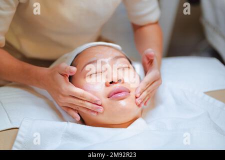 Dall'alto del raccolto non riconoscibile cosmetico fare il massaggio facciale mentre applica la maschera idratante sulla faccia del cliente asiatico femminile Foto Stock