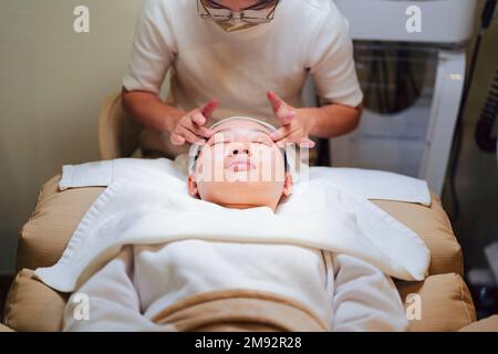 Dall'alto del raccolto non riconoscibile cosmetico fare il massaggio facciale mentre applica la maschera idratante sulla faccia del cliente asiatico femminile Foto Stock