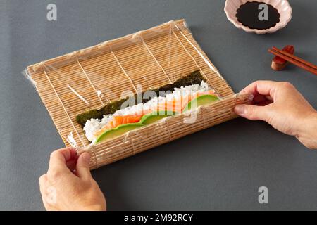 Dall'alto della persona anonima del raccolto che fa il sushi che consiste di salmone di riso di alghe e avocado con la stuoia di rotolamento di bambù sul tavolo con i chopsticks e così via Foto Stock