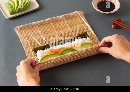 Dall'alto della persona anonima del raccolto che fa il sushi che consiste di salmone di riso di alghe e avocado con la stuoia di rotolamento di bambù sul tavolo con i chopsticks e così via Foto Stock