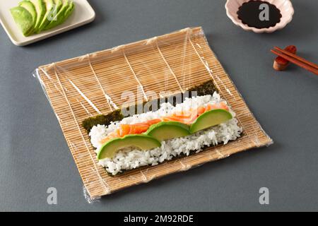 Da sopra di sushi costituito da salmone di riso di alghe e avocado posto su tappeto di rotolamento vicino a bastoncini e salsa di soia Foto Stock