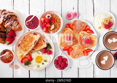Tavolo per brunch di San Valentino o Mothers Day. Vista dall'alto su uno sfondo di legno bianco. Frittelle a forma di cuore, uova e un assortimento di piatti a tema d'amore. Foto Stock
