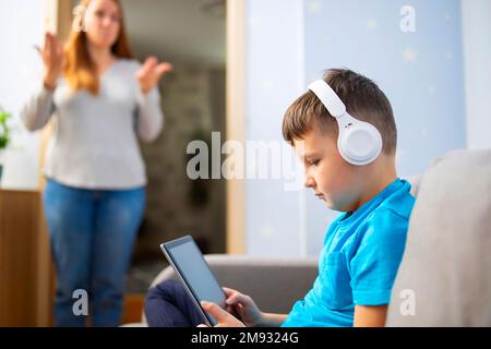 Madre scolda suo figlio. Il ragazzo usa il tablet con le cuffie e ignora la mamma Foto Stock