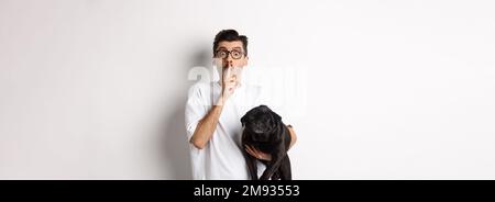 Immagine di divertente giovane uomo che regge carino cane nero pug e shushing alla macchina fotografica, mostrando il segno di hush e fissando alla macchina fotografica, mantenendo un segreto, in piedi sopra Foto Stock