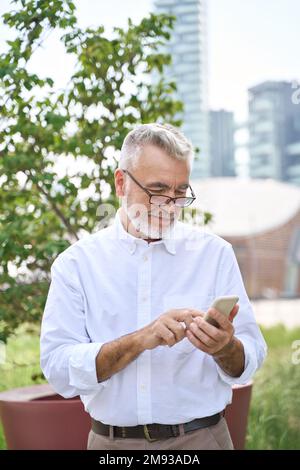 Un uomo d'affari sorridente che tiene in mano un telefono cellulare nel parco cittadino. Foto Stock