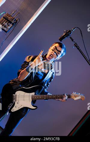 Punk rocker che fa rock sign e suona la chitarra, sentendosi estatico e facendo concerto solista in studio su sfondo. Cantante femminile che mostra il simbolo del rock and roll durante lo spettacolo dal vivo. Foto Stock