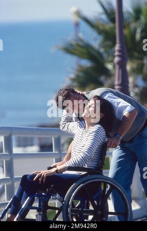 Giovane uomo che spinge una donna su una sedia a rotelle, piegandosi per darle un bacio mentre cammina fuori Foto Stock