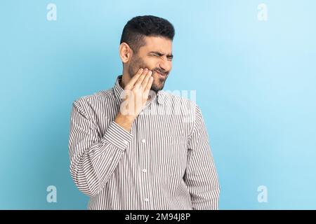 Infelice uomo d'affari sopportato sentire mal di denti, toccare guancia dolorosa, che soffrono di cavità, denti incrinati, recessione gengivale, indossare una camicia a righe. Studio in interni isolato su sfondo blu. Foto Stock