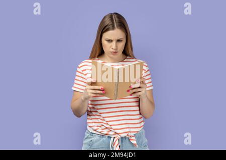 Il ritratto di una donna bionda seria e concentrata che indossa una T-shirt a righe che tiene e legge il libro, essendo impressionato dalla trama, legge con attenzione. Studio al coperto isolato su sfondo viola. Foto Stock